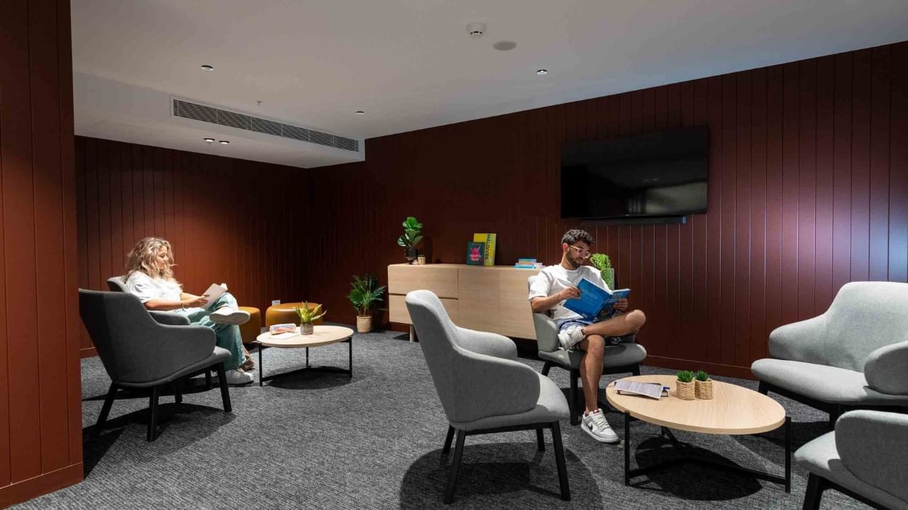 Two residents reading books in a lounge with chairs, tables, and a TV on the wall.