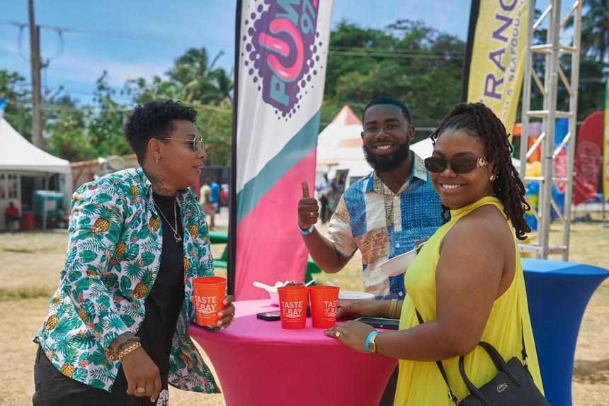 Three people chat around a colorful table, holding cups at Taste The Bay 2025 near Bay Gardens Hotels and Resorts