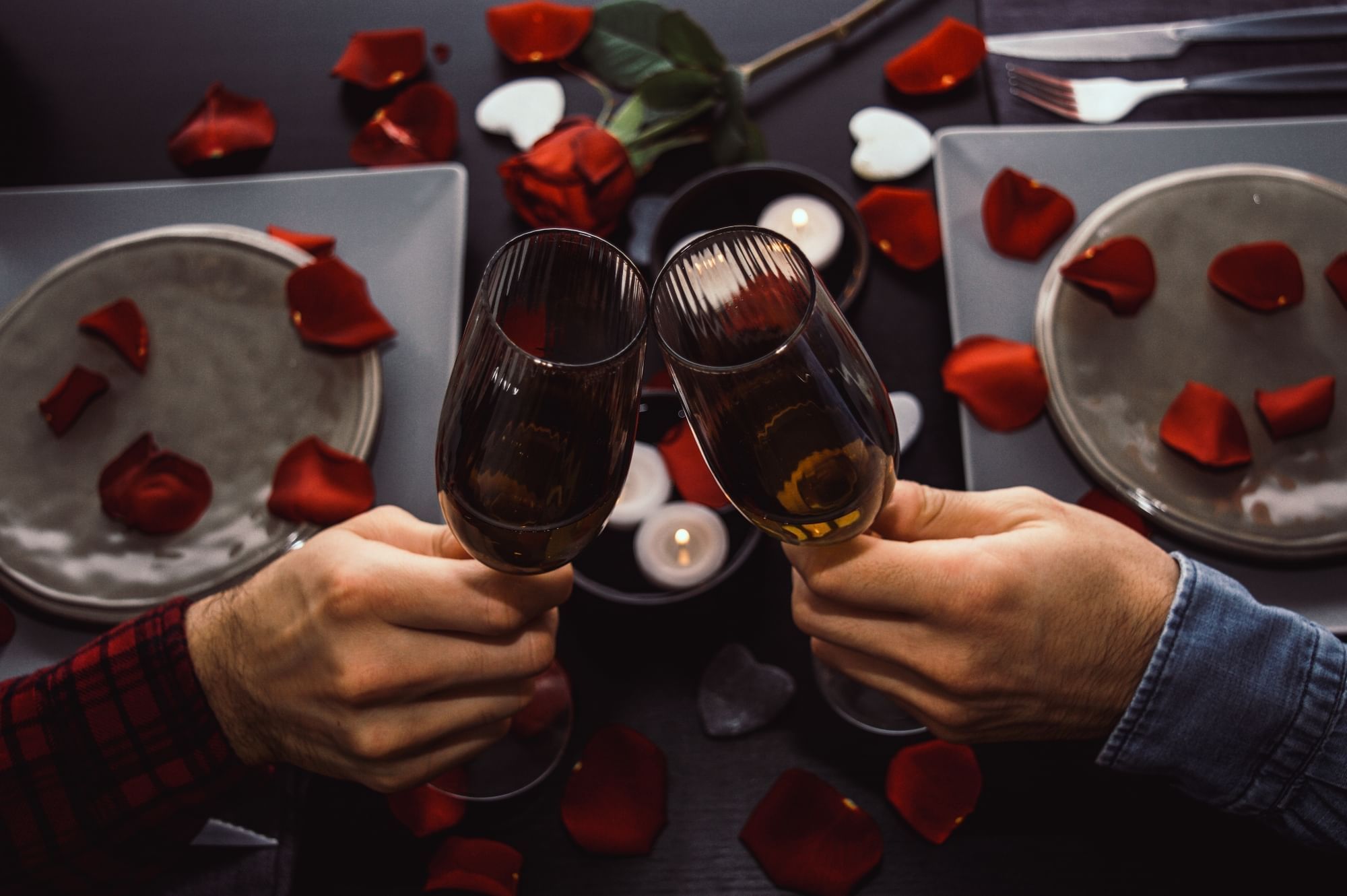 A dark table set with plates and red rose petals, with two hands holding Champagne flutes and toasting. 