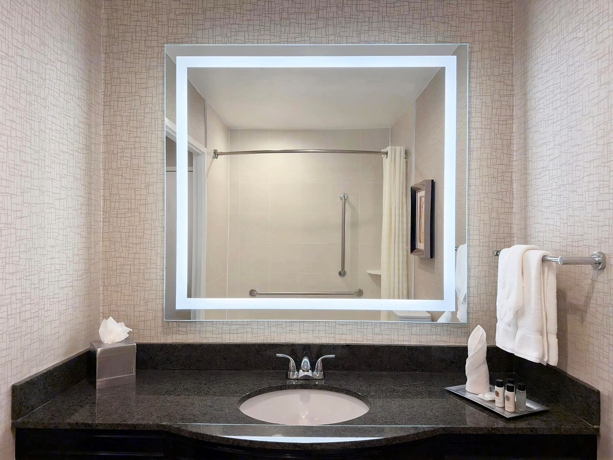 Bath sink with a mirror showing the shower area at APA Hotel Woodbridge in Iselin.