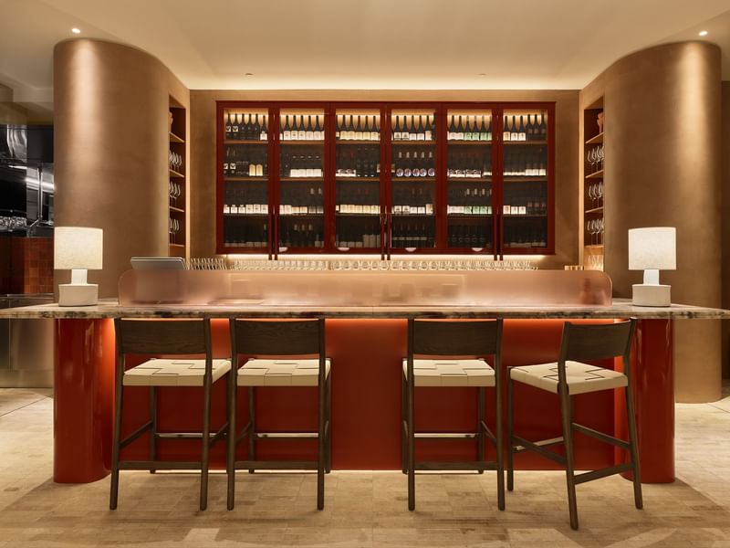 A bar counter with four stools, two lamps, and a wine cabinet with shelves and bottles.