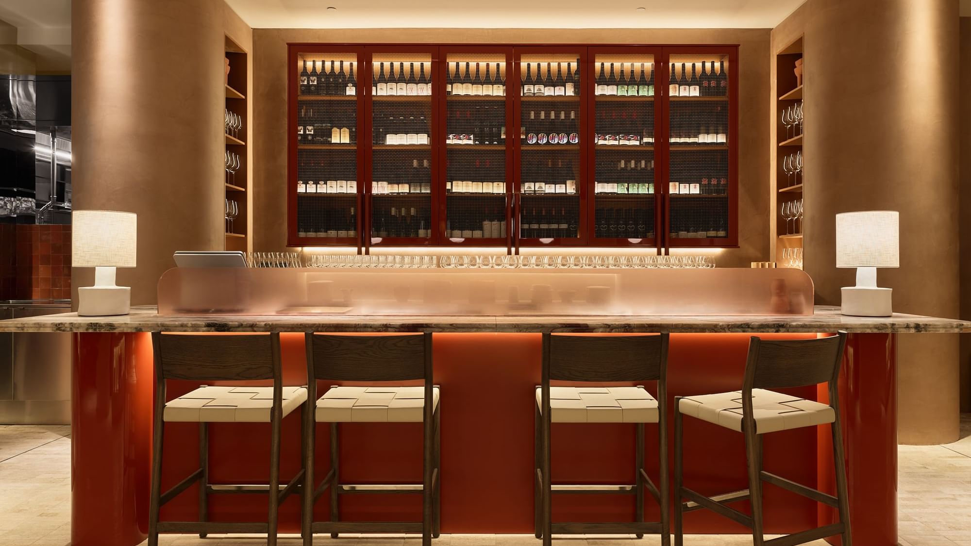A bar counter with four stools, two lamps, and a wine cabinet with shelves and bottles.
