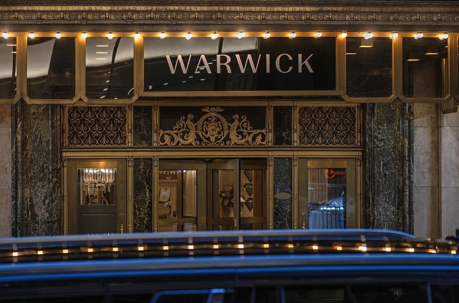 Elegant entrance of Warwick New York hotel with lit-up signage and ornate decor.