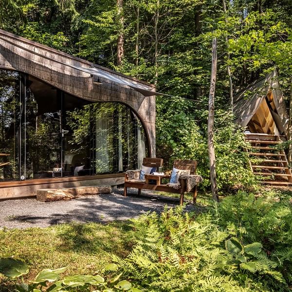 Moderne Hütte mit Glaswänden und Sitzecke im Wald neben einem zweiten Gebäude mit Treppe.