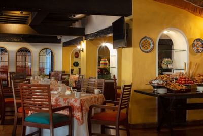 Restaurant interior with tables, chairs, yellow walls, decorative plates, and a buffet of desserts and pastries.