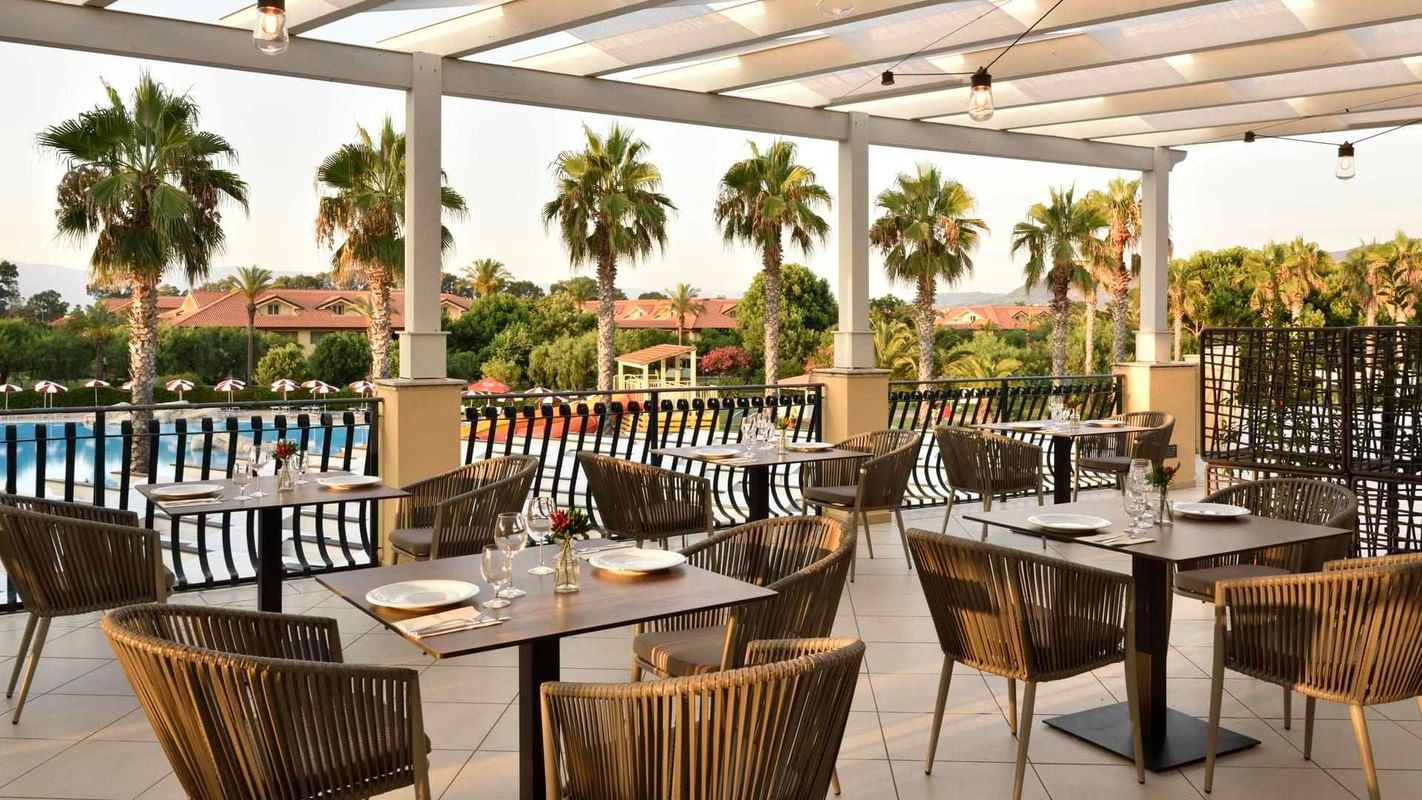 Ristorante esterno con tavoli e sedie vicino a una piscina, circondato da palme e edifici.