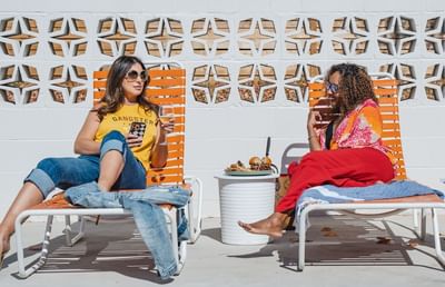 Two women relaxing on orange lounge chairs by a decorative white wall with drinks and snacks at Bardstown Motor Lodge