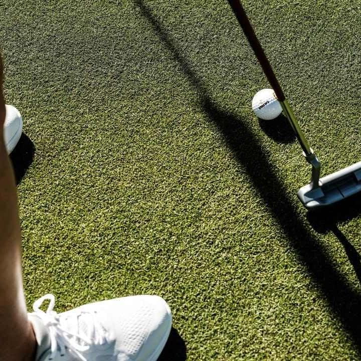 A person with white sneakers prepares to putt a golf ball with a club on green turf.