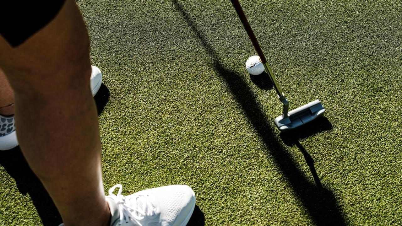 Gambe di una persona con scarpe bianche, club da golf e palla su tappeto erboso verde.