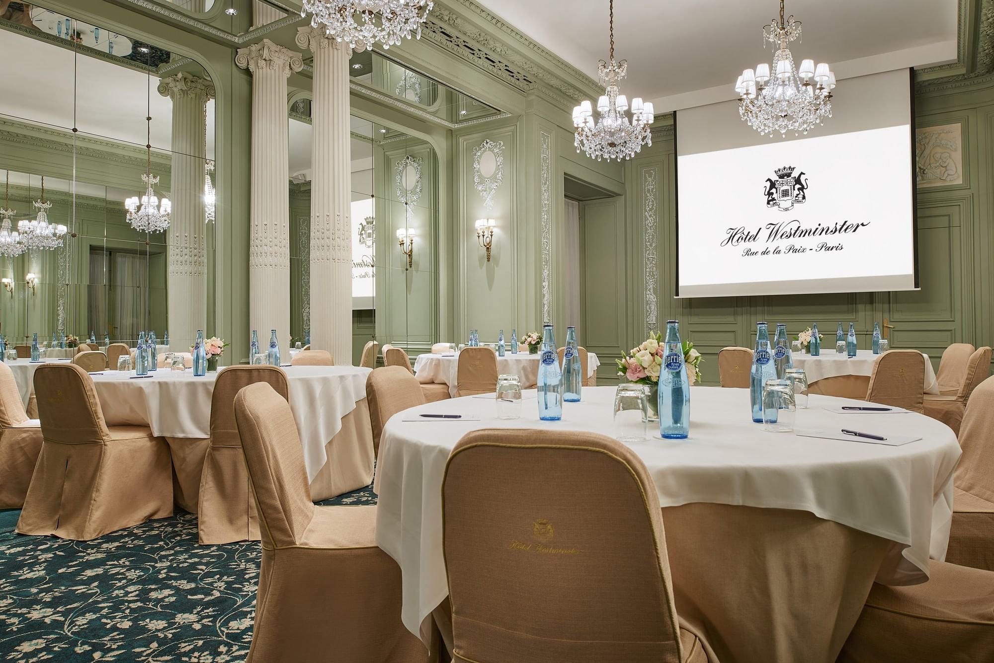 A luxurious meeting room with round tables, white tablecloths, and chairs with tan covers, set up with blue bottles, glasses, and notepads.