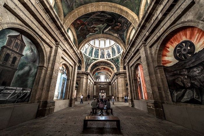Elaborate murals in a tall hall by wooden benches under a vaulted roof at Camino Real Pedregal Mexico