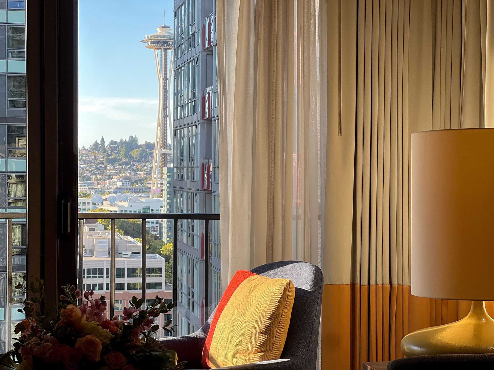 A hotel room with a view of the city, featuring a chair, lamp, and flowers.