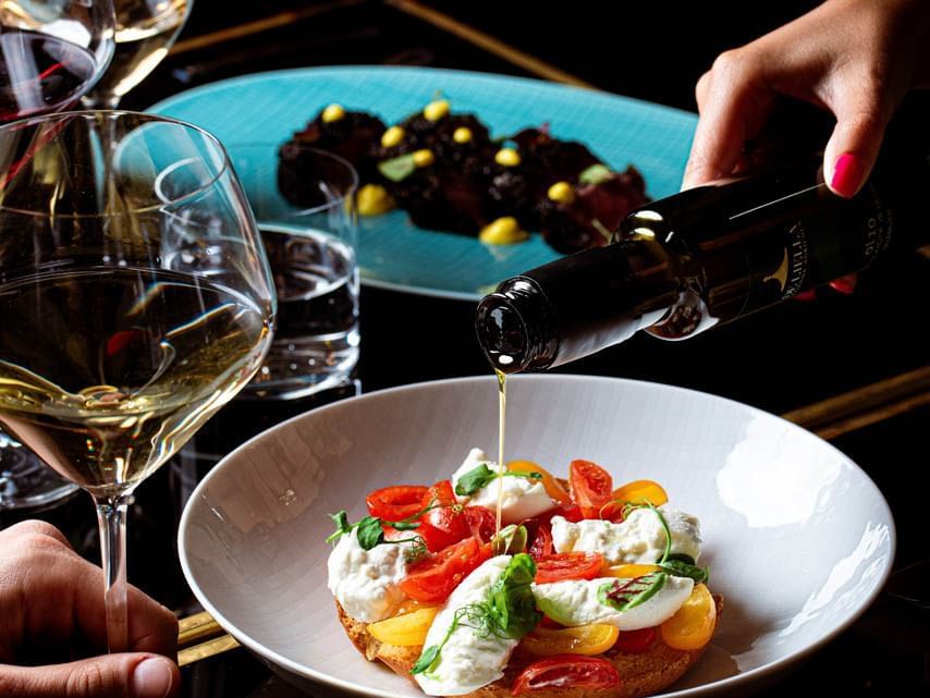 Hand drizzling olive oil on caprese salad with tomatoes and mozzarella, next to white wine at Diana’s Place, The Independent