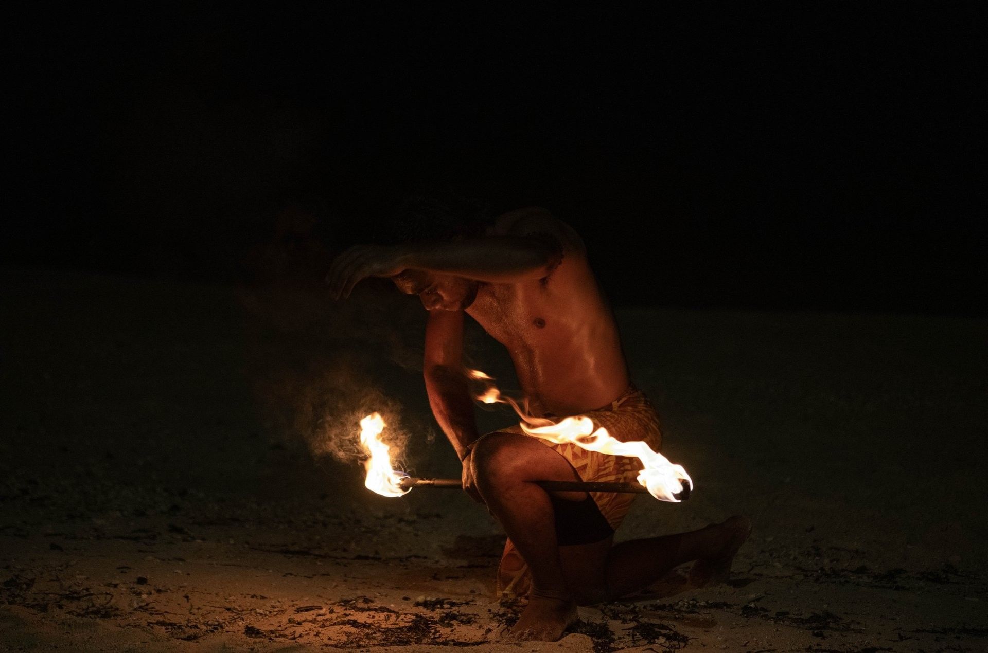 Fire dancer kneeling on a sandy beach at night under glowing embers near Warwick Fiji Resort and Spa