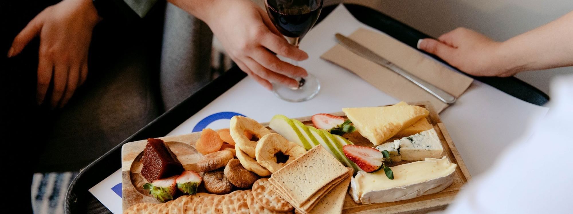 Hands holding a wine glass over a tray of assorted cheeses, crackers, fruits, and bread on a table.