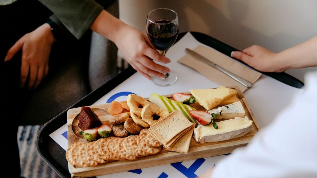 Hands holding a wine glass over a tray of assorted cheeses, crackers, fruits, and bread on a table.