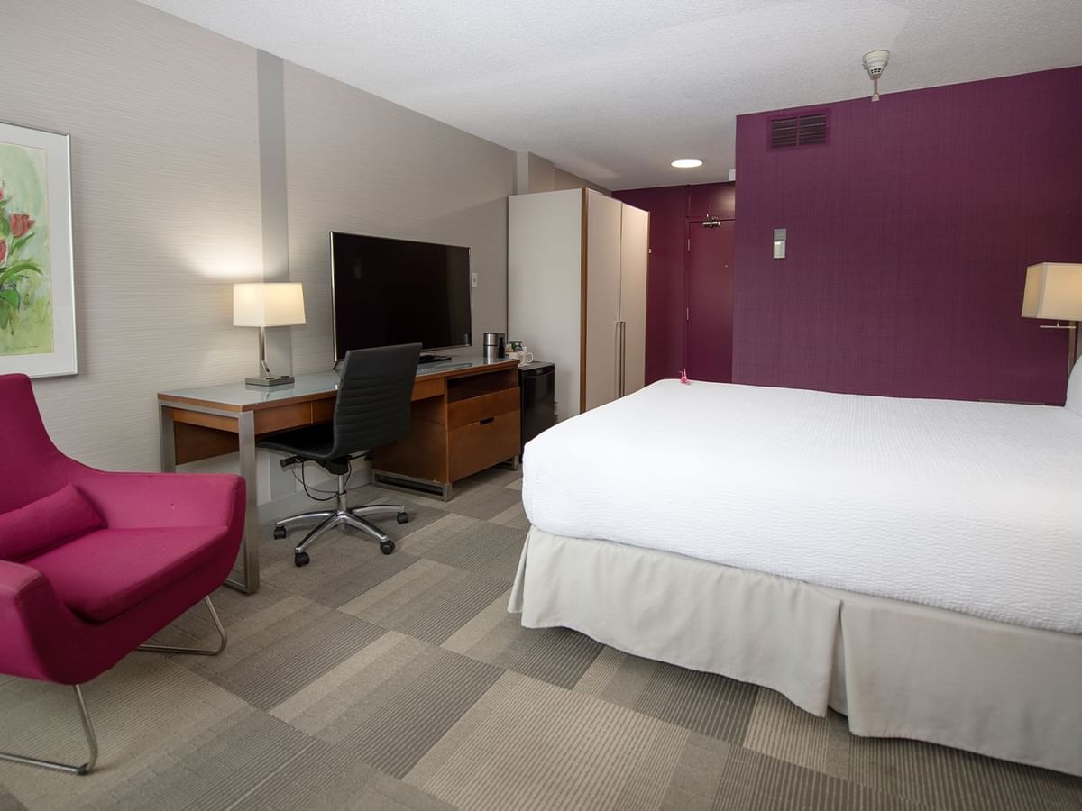 A hotel room with a bed, desk, chair, and a pink accent chair by a painting.