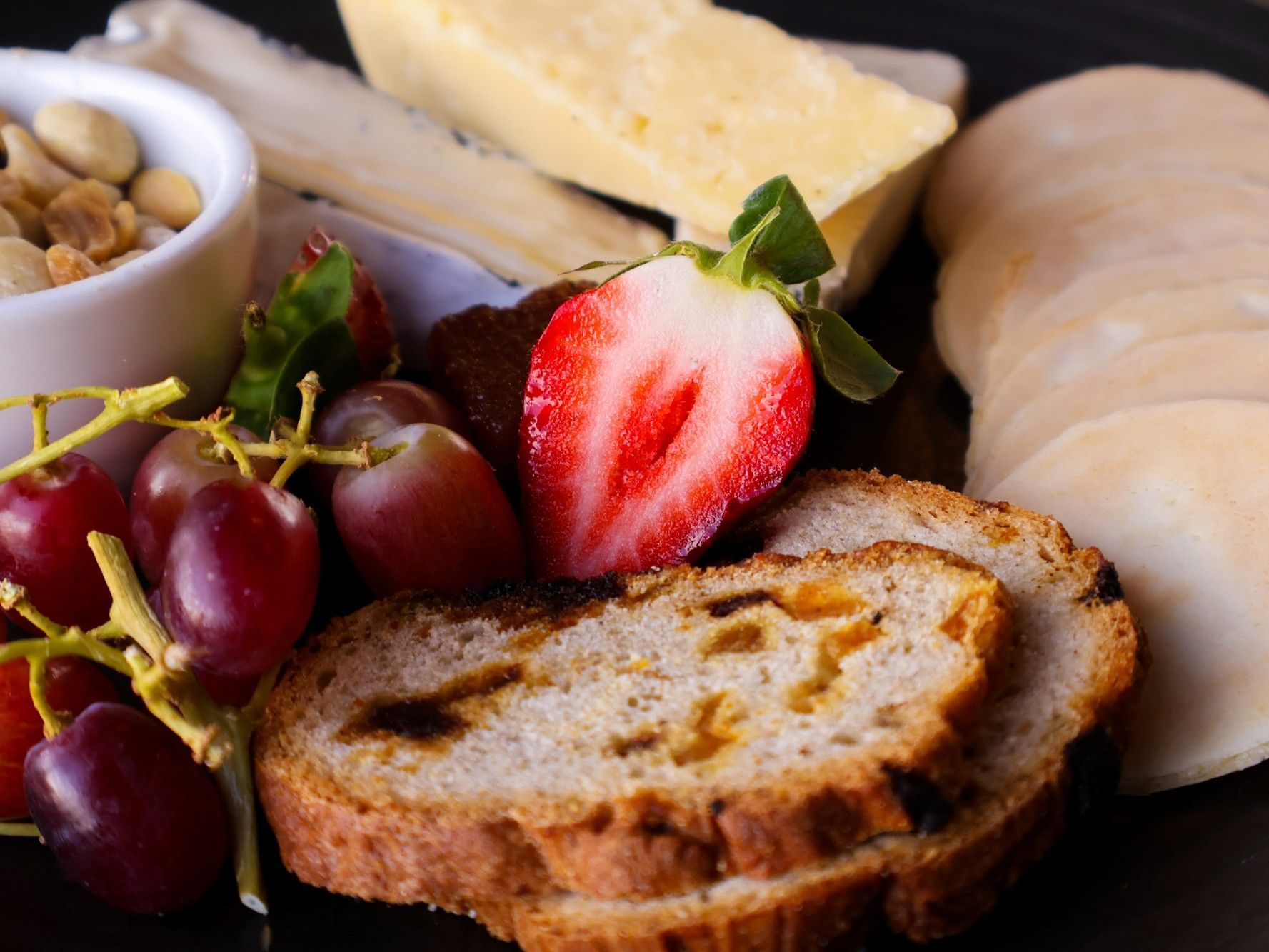 A close-up of a gourmet grazing platter featuring red grapes, a sliced strawberry, assorted cheeses, thin crackers, toasted fruit bread, and a small bowl of nuts