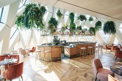 Interior of a bar featuring plush seating and abundant greenery against large windows at Hotel Motto Vienna