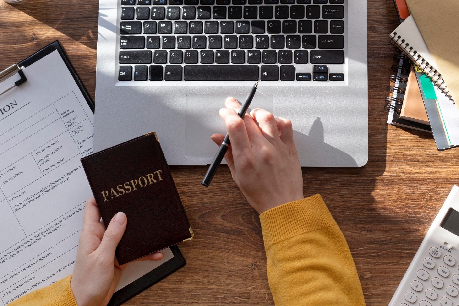 Person holding a passport while filling out travel documents on a laptop, with visa application form, calculator, and notebooks on a wooden desk, symbolizing international travel planning and preparation.
