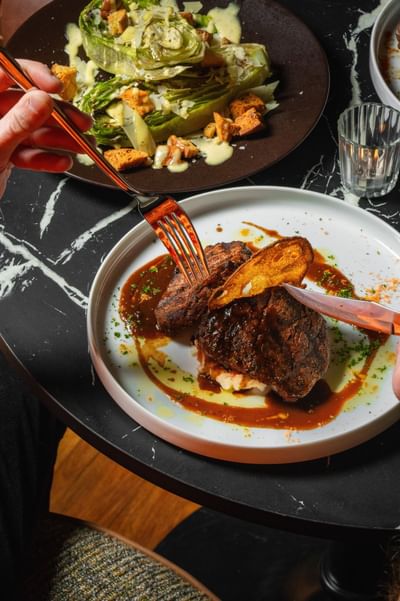 Hand using fork & knife to cut into a rich beef dish with sauce on a white plate at Cypress & Oak steakhouse in Frankfort, KY