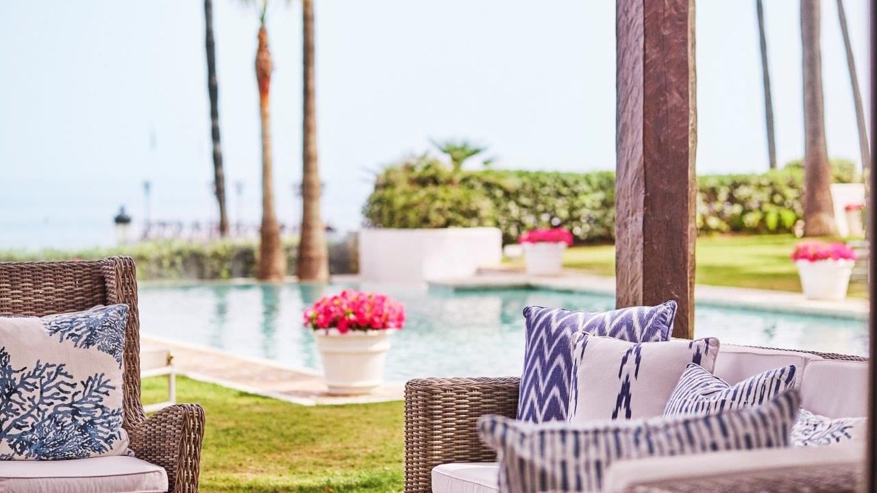 Villa del Mar with poolside wicker lounge seating with patterned cushions, palm trees, & pink flowers at Marbella Club