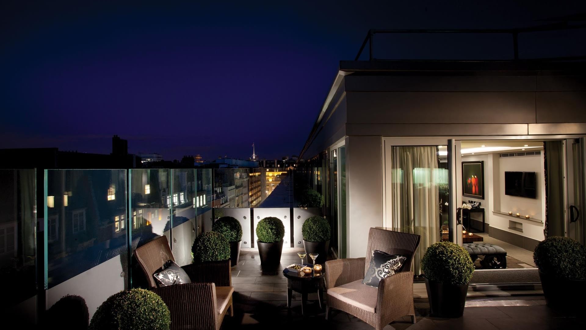 Stylish rooftop terrace at night with wicker chairs and small round tables  in The Ebony Suite at The May Fair Hotel London