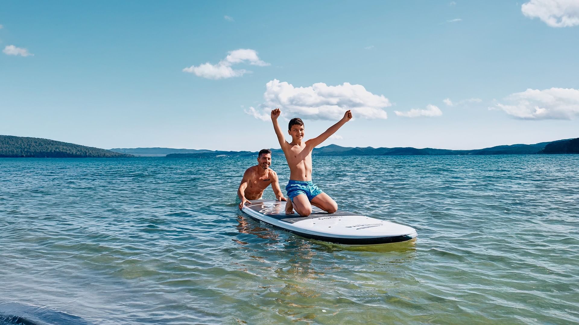 Padre e figlio in piedi su una tavola da paddle-board nel mare con le braccia alzate al cielo da Falkensteiner Group