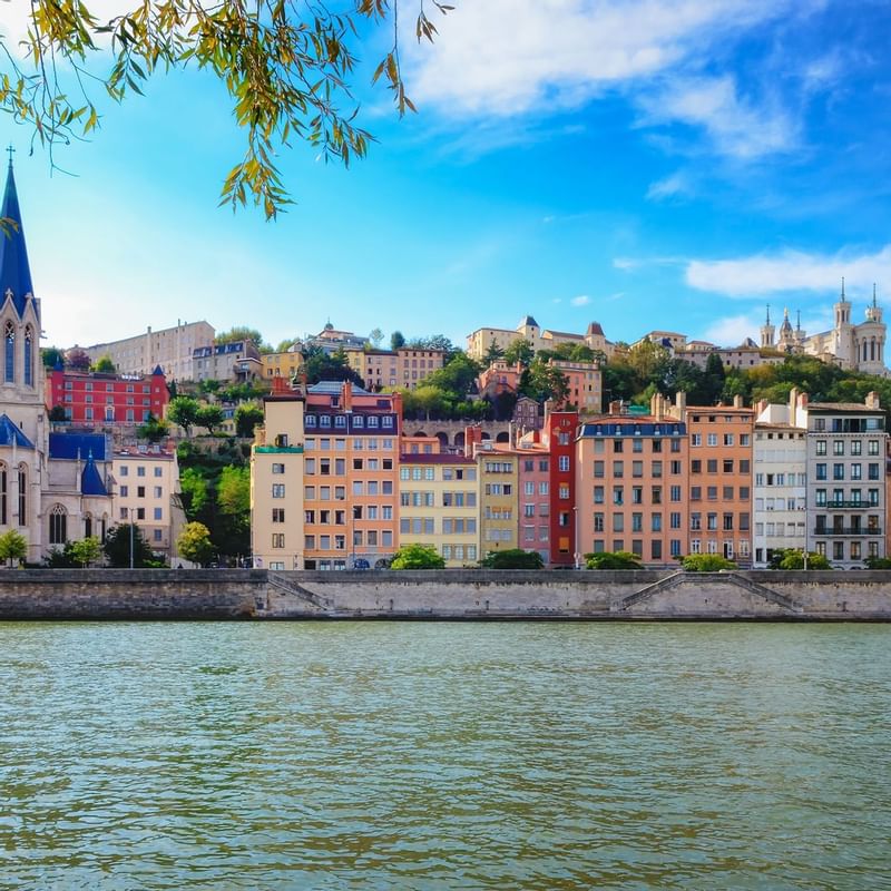 Colorful buildings line the riverside in Lyon, with a prominent church spire, hills and Warwick Hotels and Resorts