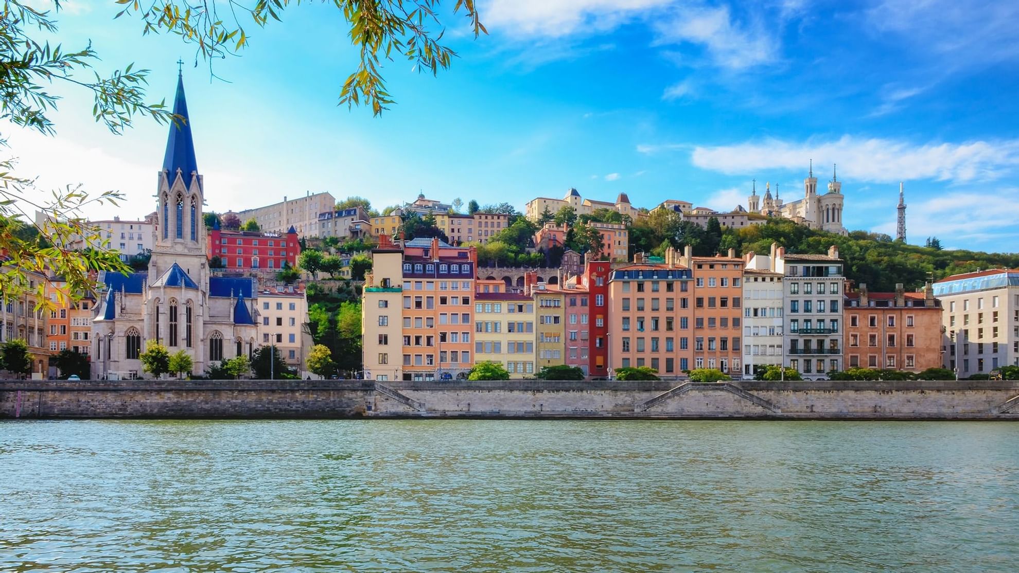 Colorful buildings line the riverside in Lyon, with a prominent church spire, hills and Warwick Hotels and Resorts