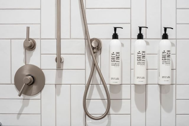 A shower with white tiles, a silver shower head and controls, and three bottles of toiletries.