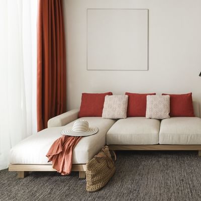 Soggiorno minimalista con divano beige, cuscini rossi, tavolo, sedie, borsa e cappello.