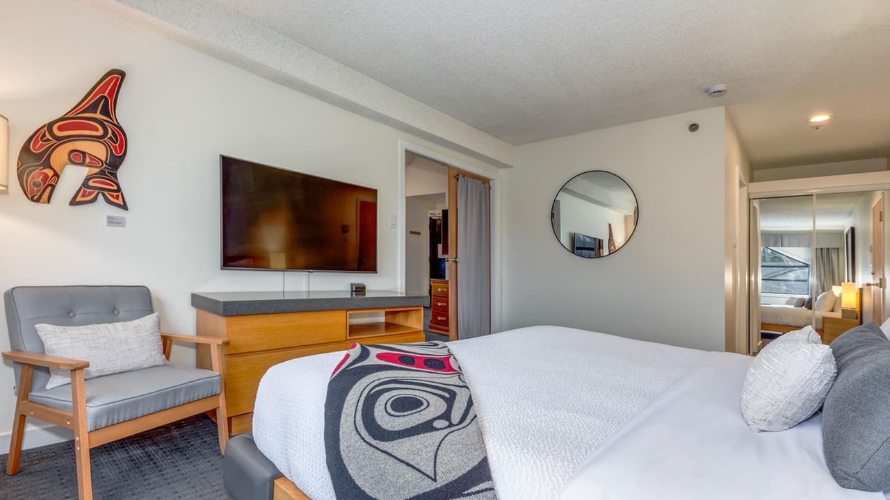 A hotel room with a bed, chair, TV, dresser, mirror, and indigenous art on the wall.