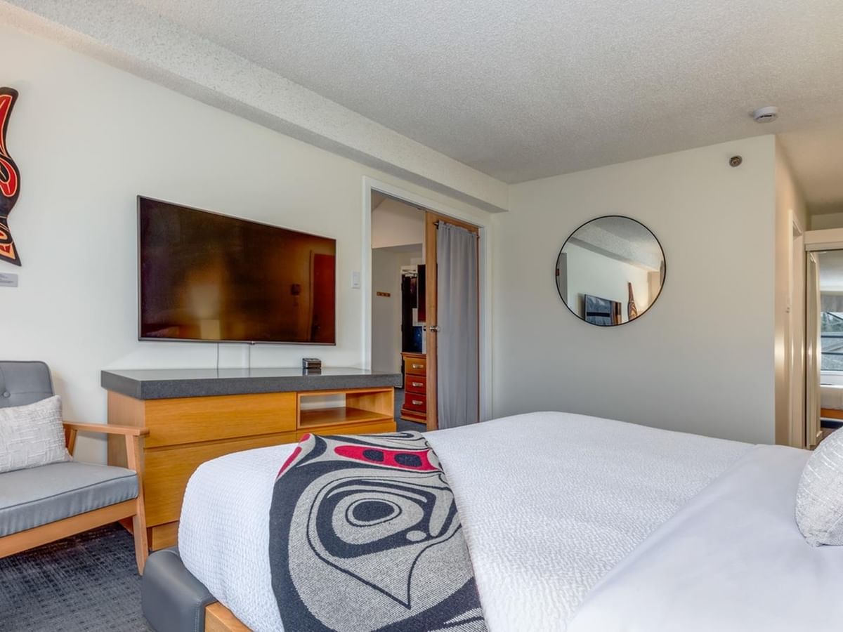 A bedroom with a bed, chair, TV, dresser, mirror, and Indigenous art on the wall.