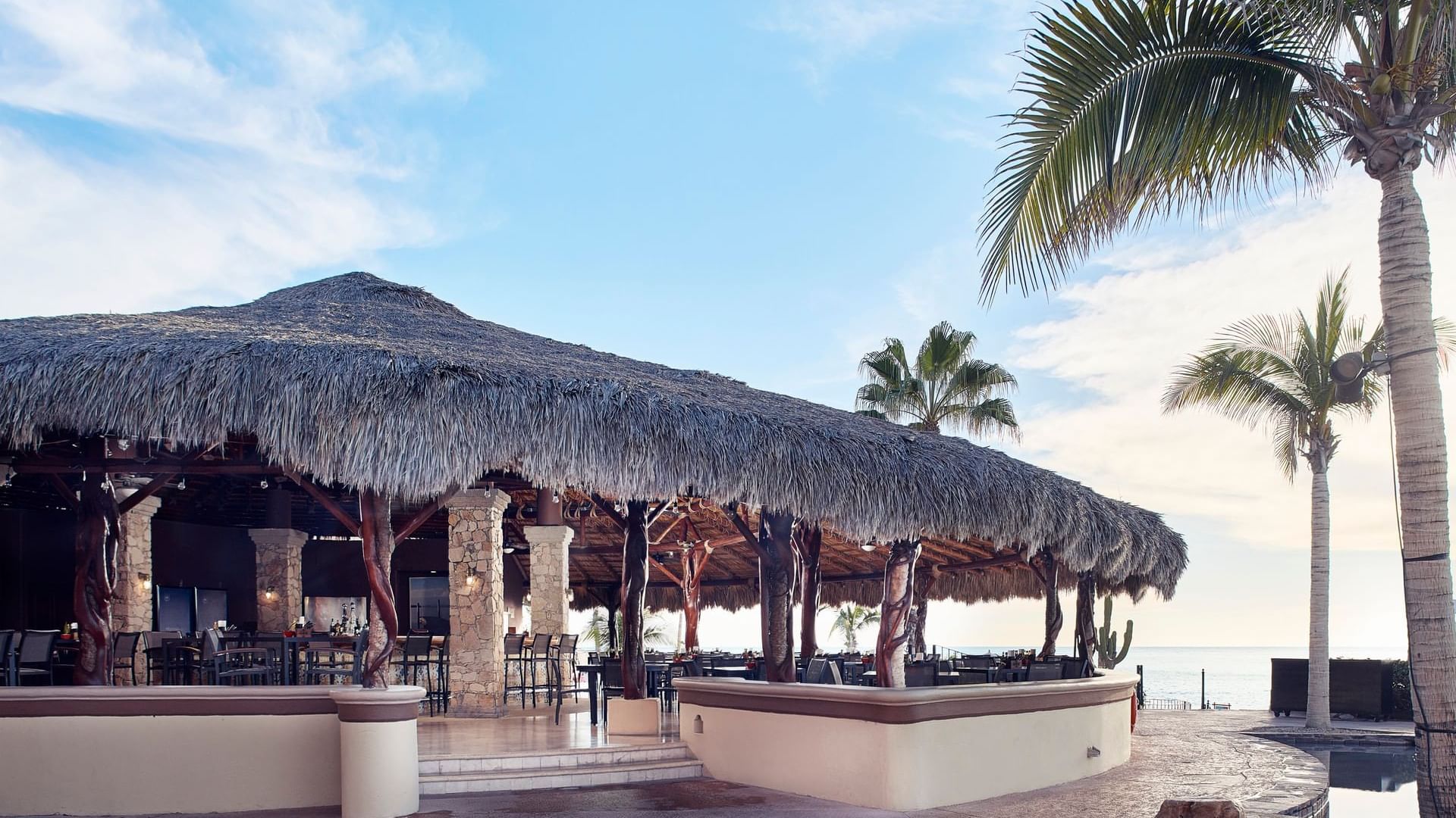 Sirenas Restaurant's exterior with a thatched roof, bar stools, and ocean views at Hacienda del Mar Los Cabos.