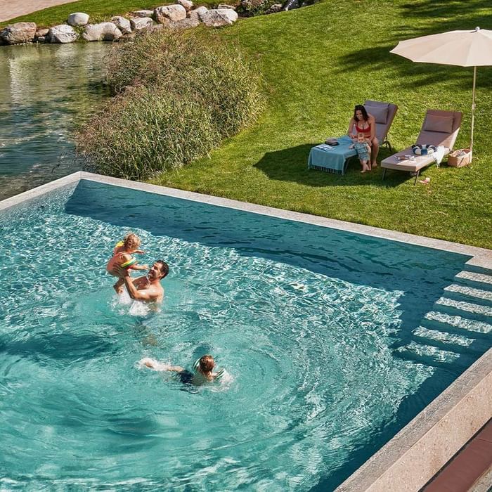 Famiglia di tre persone in piscina, con due genitori e due bambini, uno seduto sulla spiaggia.
