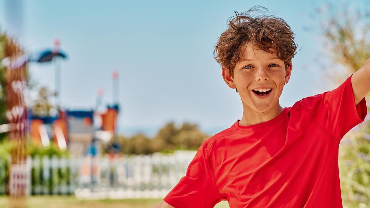 Lächelnder Junge in rotem T-Shirt vor Spielplatz mit weißen Holzzäunen.
