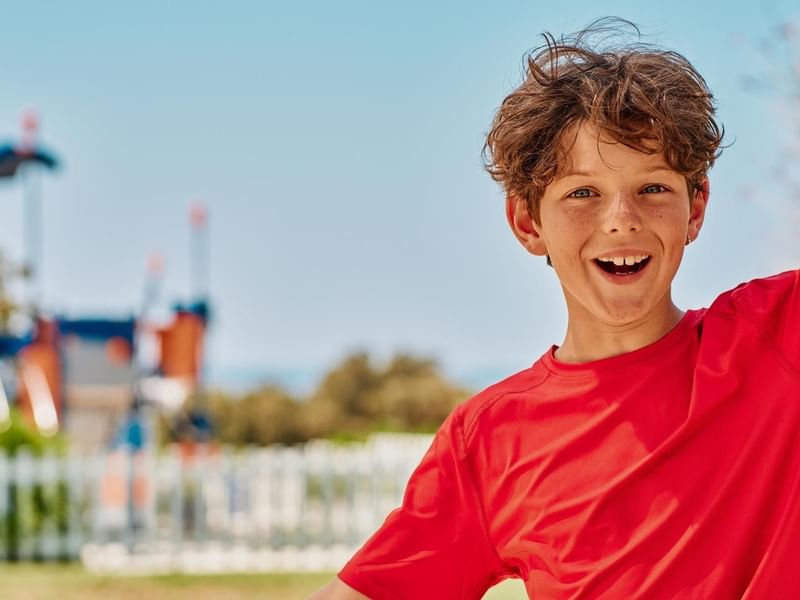 Lächelnder Junge in rotem T-Shirt vor Spielplatz mit weißen Holzzäunen.