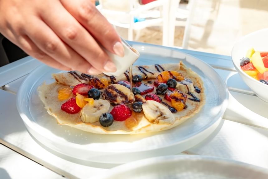 Hand pouring syrup on a pancake topped with various fruits and chocolate drizzles on a white plate.