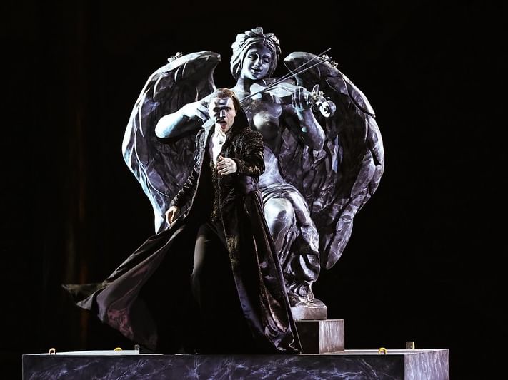 Man in black cloak stands on stage in front of large angel statue holding a bow and violin.
