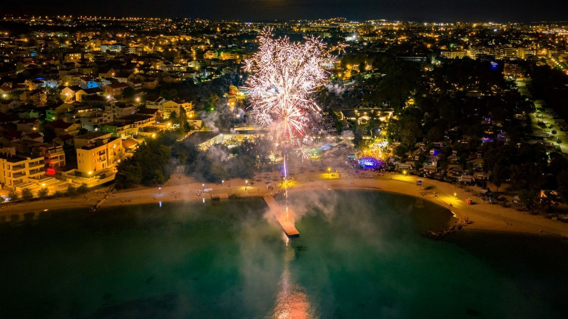 Fuochi d'artificio notturni sopra una città costiera illuminata e una spiaggia affollata.