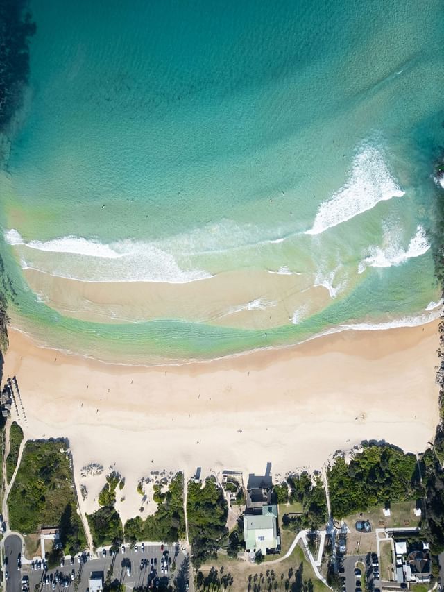 Aerial view of waves rolling onto Freshwater Beach sand, surrounding green headlands near Elysium Manly, best hotels in Manly