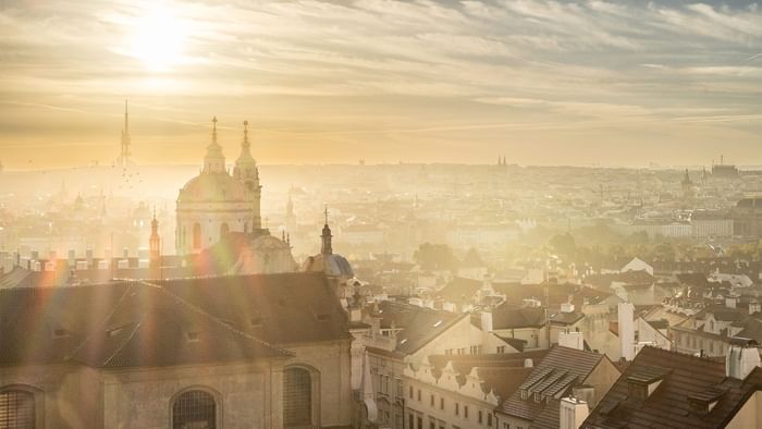 Sunrise over a misty cityscape with historic buildings and church domes under sunlight near Falkensteiner Hotel Prague