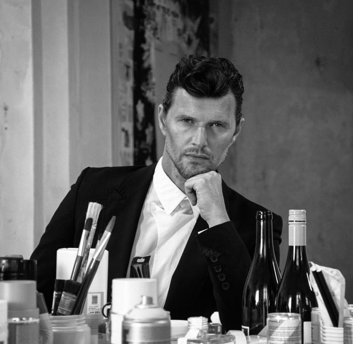 Sasha Knezevic sitting at a cluttered table filled with art supplies, paint cans, and bottles at Hotel Motto Vienna