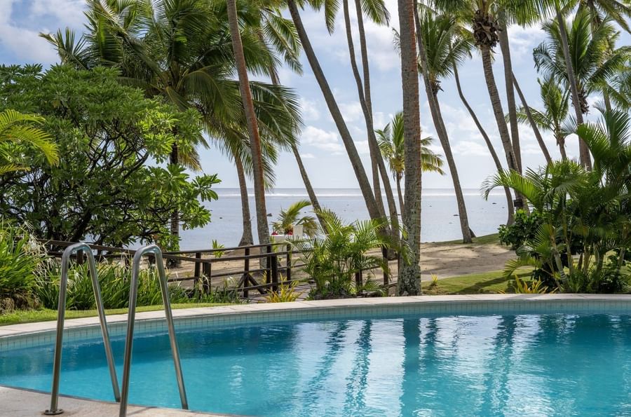 Blue swimming pool by a dark wooden fence under tall palm trees near the sandy shore at Tambua Sands Beach Resort