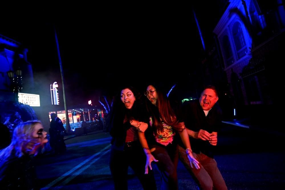 Two women and a man scream and cling to each other in terror as a zombie approaches them on a dark street.
