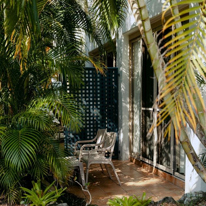 Two white chairs and a table beside a black lattice door on a tiled patio with tropical plants.