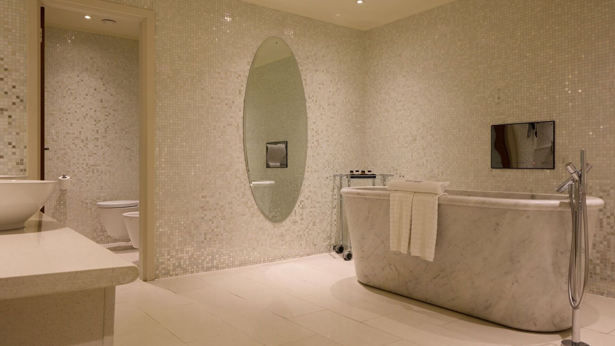 Spacious bathroom with mosaic tile walls, freestanding tub, and oval mirror in The Penthouse Suite at The May Fair Hotel, London.