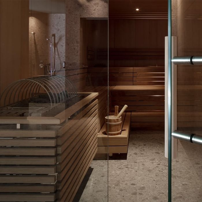 Innenansicht einer Sauna mit Holzbänken, einem Eimer und einem Glastür.