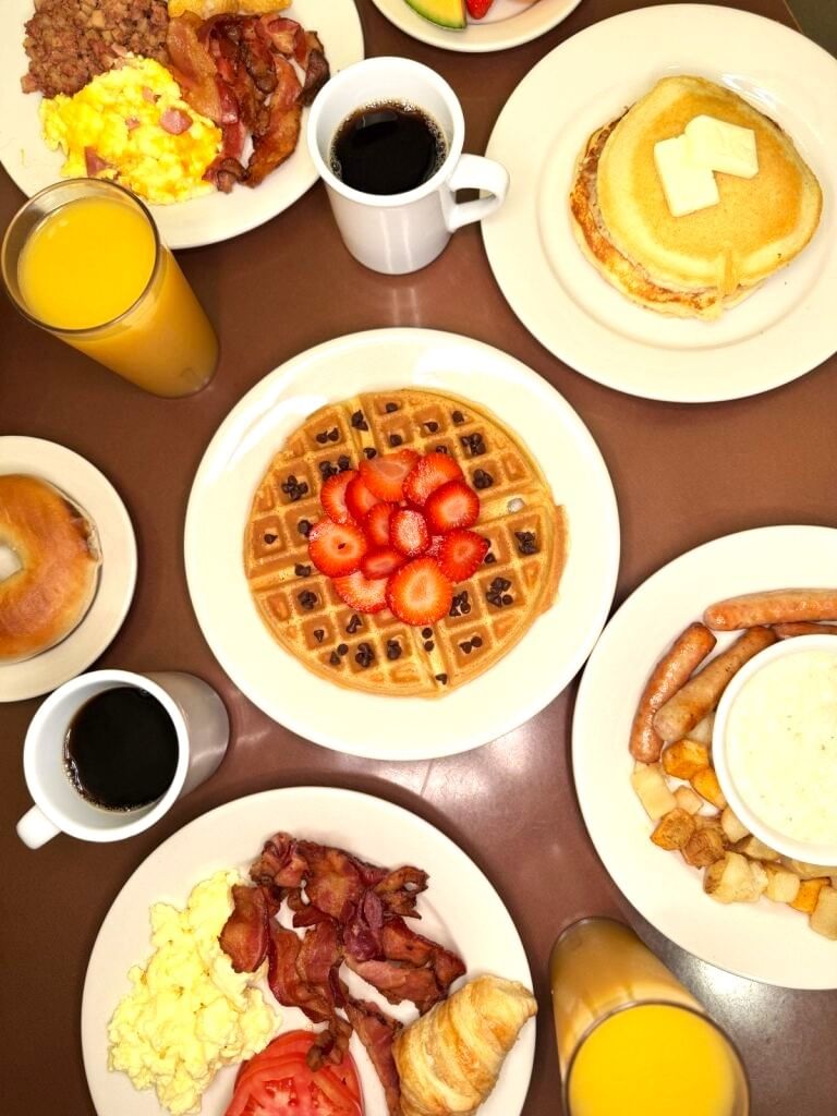 An overhead shot of a table set with breakfast food, including a waffle with strawberries, pancakes, bacon, a bagel, sausage, and cups of orange juice and coffee.